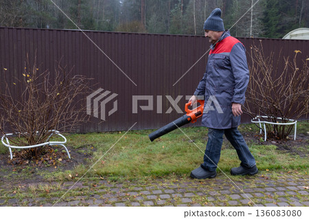 Worker removing fallen leaves with electric blower, damp weather, everyday maintenance task, suburban environment, realistic lifestyle scene.  136083080