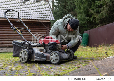 Man inspects red lawnmower near wooden house wearing puffy jacket beanie crouched in outdoor setting. Man inspects red lawnmower near wooden house wearing puffy jacket beanie crouched in outdoor setting. 136083082