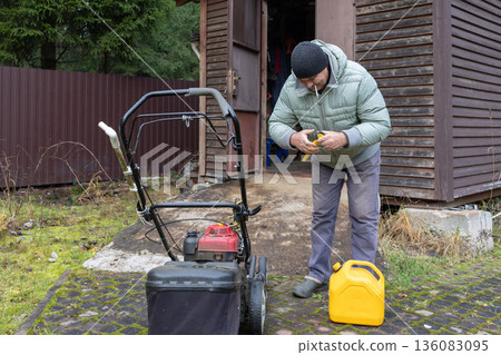 Man in wearing warm jacket and beanie fills lawnmower fuel tank with yellow can near shed. Overcast day outdoor maintenance. Man in wearing warm jacket and beanie fills lawnmower fuel tank with yellow can near shed. Overcast day outdoor maintenance. 136083095