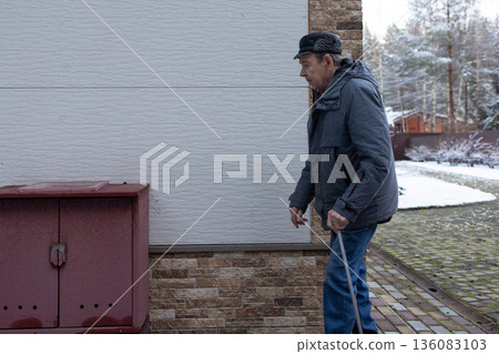 Senior male moving carefully with walking stick on brick pavement surrounded by snow covered landscape cold season daily activity. 136083103