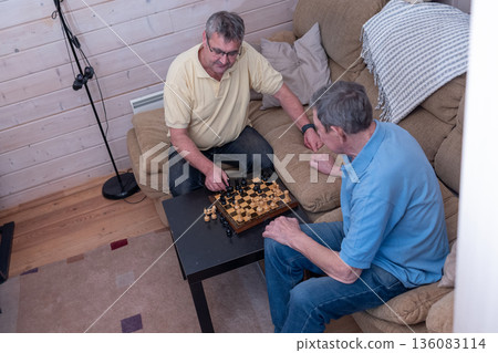 Elderly friends concentrating on chess match in living room in casual clothing. Thoughtful mood and indoor recreation scene. Elderly friends concentrating on chess match in living room in casual clothing. Thoughtful mood and indoor recreation scene. 136083114