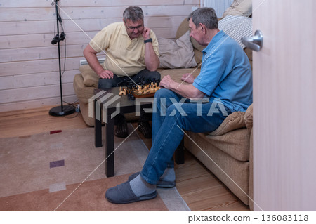 Elderly friends concentrating on chess match in living room in casual clothing. Thoughtful mood and indoor recreation scene.  136083118