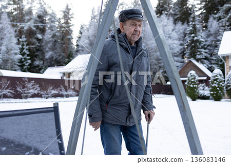 Elderly man walking with cane on paved yard after snowfall wearing winter jacket and hat outdoors calm winter environment. Elderly man walking with cane on paved yard after snowfall wearing winter jacket and hat outdoors calm winter environment. 136083146