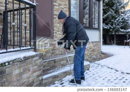 Adult man in winter clothing sweeping snow from porch steps with broom, outdoor home area in cold season maintenance task. Adult man in winter clothing sweeping snow from porch steps with broom, outdoor home area in cold season maintenance task. 136083147