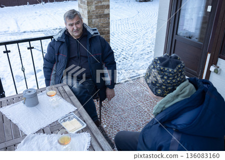 Two senior men sitting at table on terrace in winter, calm dialogue, hot drinks, everyday lifestyle and social connection Two senior men sitting at table on terrace in winter, calm dialogue, hot drinks, everyday lifestyle and social connection 136083160