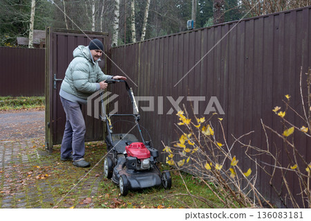 Man mows lawn with red lawnmower in front of house, greenery around, overcast day. Garden maintenance task in overcast day 136083181