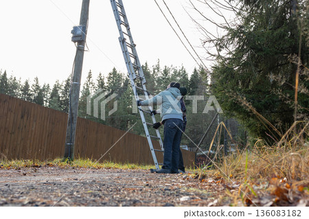 Two adult men using ladder at utility pole when replacing lamp on rural road surrounded by trees and houses during autumn daylight. 136083182