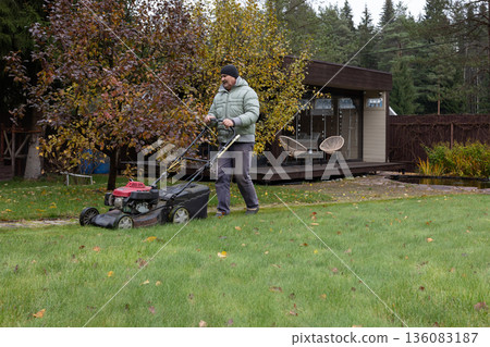 Man uses lawnmower to trim grass in front of residential home and lush greenery. Calm outdoor setting. Man uses lawnmower to trim grass in front of residential home and lush greenery. Calm outdoor setting. 136083187