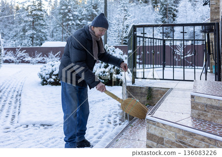 Adult man in winter clothing sweeping snow from porch steps with broom, outdoor home area in cold season maintenance task. 136083226