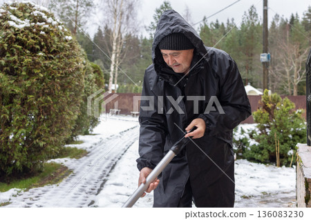 Elderly man in dark winter clothing clearing snow with shovel on paved yard area emphasizing seasonal outdoor work Elderly man in dark winter clothing clearing snow with shovel on paved yard area emphasizing seasonal outdoor work 136083230