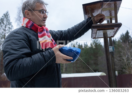 Man refilling bird feeder with seeds in snowy garden wearing jacket and scarf among trees during cold season. 136083263