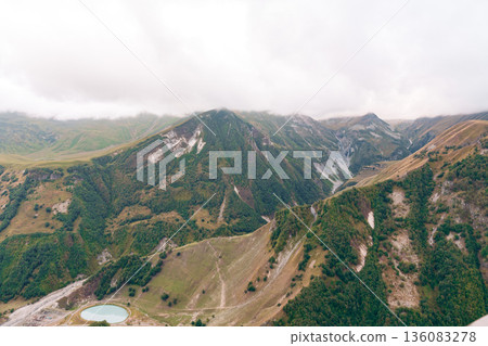 View of mountains with green slopes and a lake under cloudy sky in a natural landscape View of mountains with green slopes and a lake under cloudy sky in a natural landscape 136083278