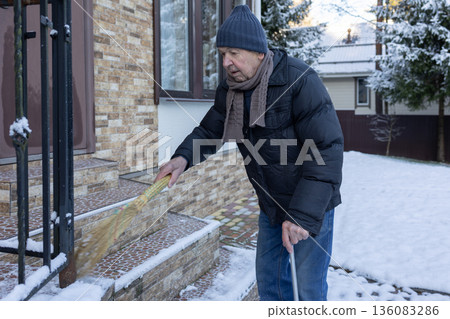 Adult man in winter clothing sweeping snow from porch steps with broom, outdoor home area in cold season maintenance task. Adult man in winter clothing sweeping snow from porch steps with broom, outdoor home area in cold season maintenance task. 136083286