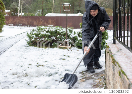 Elderly man in dark winter clothing clearing snow with shovel on paved yard area emphasizing seasonal outdoor work 136083292