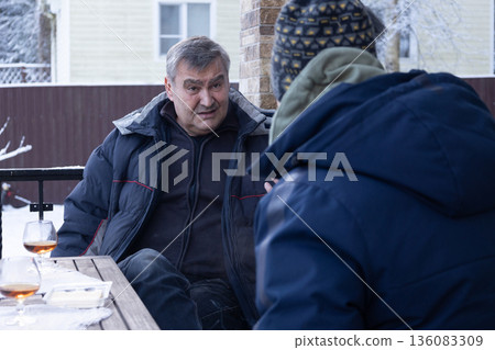 Two senior men sitting at table on terrace in winter, calm dialogue, hot drinks, everyday lifestyle and social connection 136083309