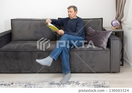Elderly man sitting on sofa reading book in calm living room environment emphasizing leisure and quiet indoor lifestyle. Elderly man sitting on sofa reading book in calm living room environment emphasizing leisure and quiet indoor lifestyle. 136083324
