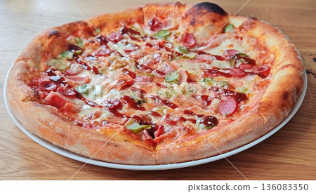 Freshly Baked Pepperoni and Ham Pizza Closeup. Delicious homemade pizza topped with pepperoni, ham, pickles, veggies, and sauce on wooden table. 136083350