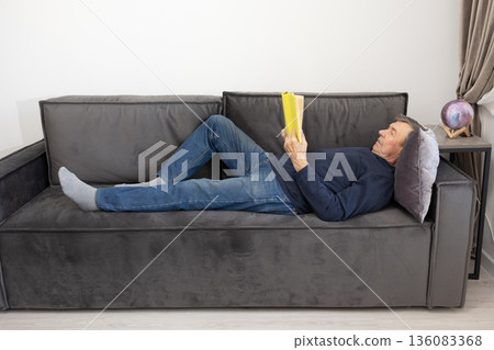 Elderly man lying on sofa reading book in calm living room environment emphasizing leisure and quiet indoor lifestyle. Elderly man lying on sofa reading book in calm living room environment emphasizing leisure and quiet indoor lifestyle. 136083368