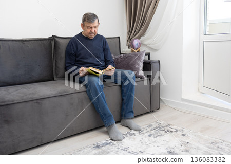 Elderly man sitting on sofa reading book in calm living room environment emphasizing leisure and quiet indoor lifestyle. 136083382