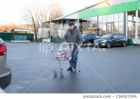 Elderly man in outdoor clothing standing on parking lot with shopping cart full of groceries near personal vehicle 136083384