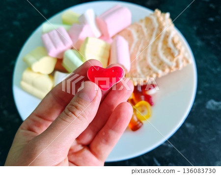 Hand Holding Red Heart Candy with Sweets Assortment. Female hand holding heart-shaped red gummy candy over plate of colorful marshmallows and sweets. 136083730