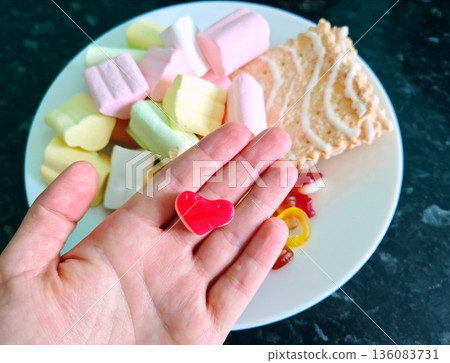 Hand Holding Red Heart Candy with Sweets Assortment. Female hand holding heart-shaped red gummy candy over plate of colorful marshmallows and sweets. 136083731