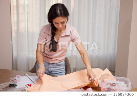 Woman measuring light orange fabric with a tape measure on a wooden table, surrounded by sewing supplies 136084300