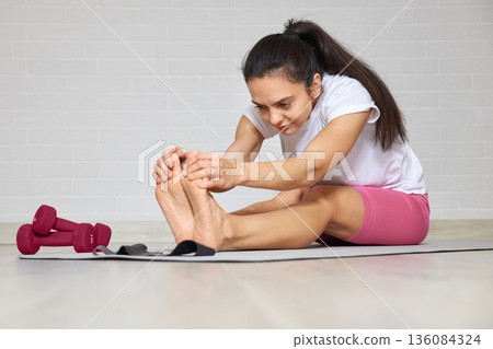 Woman performing a seated forward bend on a yoga mat with dumbbells nearby 136084324