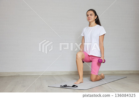 Young woman training making lunges with a dumbbell on a mat indoors 136084325