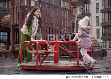 Mother and daughter playing together on a merry-go-round in an urban park Mother and daughter playing together on a merry-go-round in an urban park 136084334
