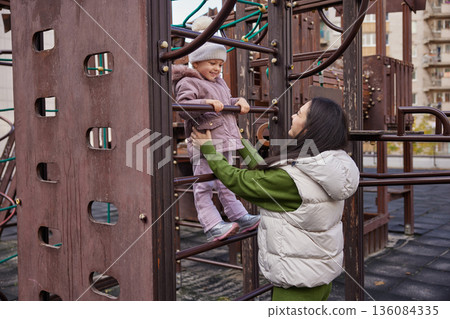 Mother assisting child on monkey bars at a playground, sharing a happy moment 136084335