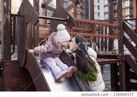 Mother and daughter touching noses, playing together on a playground slide 136084337