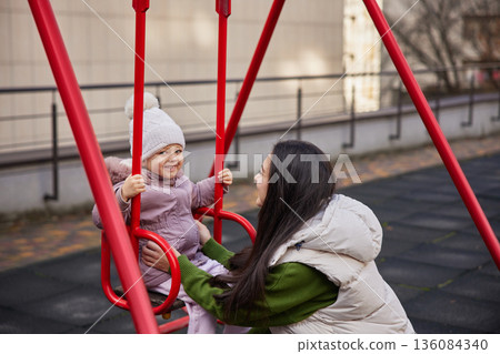 Mother pushing happy daughter on a swing set in an urban playground Mother pushing happy daughter on a swing set in an urban playground 136084340