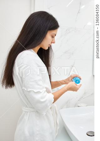 Woman in bathrobe dispensing lotion from a blue bottle into her hand in the bathroom 136084350