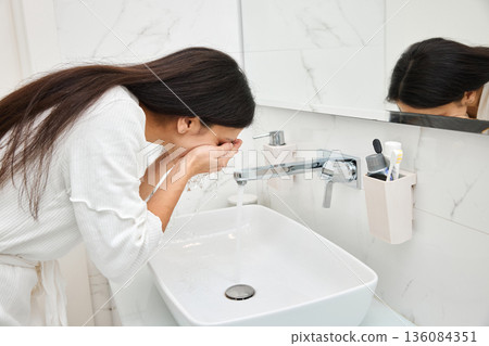 Woman washing her face with water from a modern faucet in a white bathroom 136084351