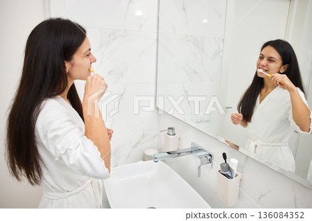Woman brushing teeth in bathroom mirror, focusing on oral hygiene as part of her morning routine 136084352
