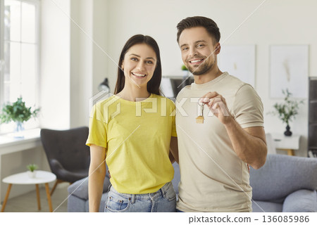 Happy family couple standing together in new modern living room holding flat key, moving  136085986