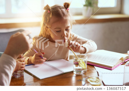 Little girl drinking water with lemon while drawing at table. Little girl drinking water with lemon while drawing at table. 136087317