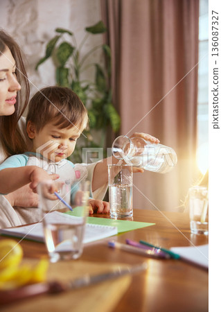 Mother pouring water into glass at table during family activity. 136087327