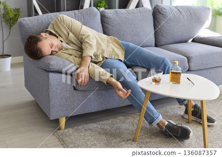 Young man passed out after heavy drinking, lying on couch with whiskey bottle 136087357