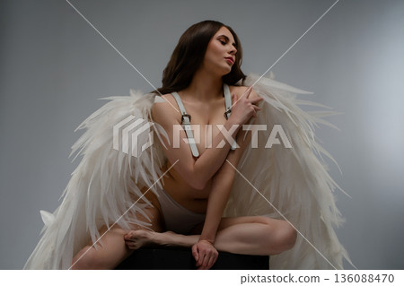 Ethereal woman with long brown hair wearing large white angel wings and a harness, posing in a studio. 136088470