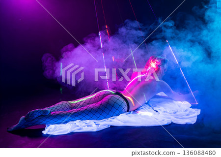 Sensual woman in fishnet stockings lying in a futuristic club setting with neon blue and pink lights, smoke, and red laser beams. Sensual woman in fishnet stockings lying in a futuristic club setting with neon blue and pink lights, smoke, and red laser beams. 136088480