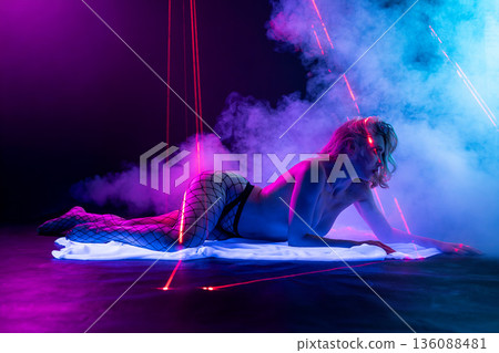 Sensual woman in fishnet stockings lying in a futuristic club setting with neon blue and pink lights, smoke, and red laser beams. Sensual woman in fishnet stockings lying in a futuristic club setting with neon blue and pink lights, smoke, and red laser beams. 136088481