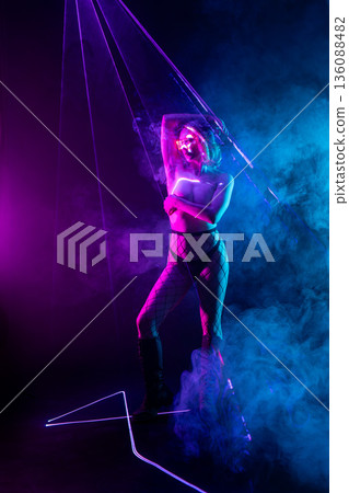 Cyberpunk woman in futuristic glasses stands amidst neon purple laser beams and blue smoke. Cyberpunk woman in futuristic glasses stands amidst neon purple laser beams and blue smoke. 136088482