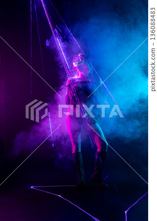 Cyberpunk woman in futuristic glasses stands amidst neon purple laser beams and blue smoke. 136088483