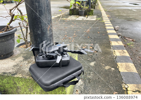 compact foldable drone and remote control resting on a black travel case outdoors on a damp concrete ledge, ready for flight 136088974