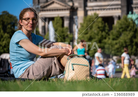 Berlin, germany, august 11, 2023. Woman sitting on the green grass, relaxing and looking at the camera, with the berlin cathedral in the blurry background 136088989