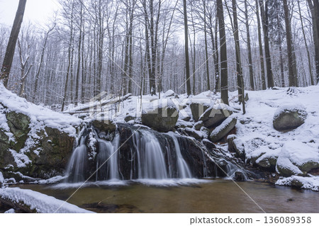 Winter forest waterfall flowing through snow covered rocks Winter forest waterfall flowing through snow covered rocks 136089358