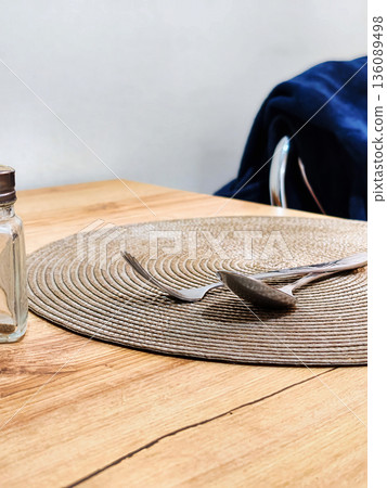 A round placemat is placed on a wooden table. A spoon and a fork are resting on the placemat next to a small glass jar. A chair is in the background A round placemat is placed on a wooden table. A spoon and a fork are resting on the placemat next to a small glass jar. A chair is in the background 136089498