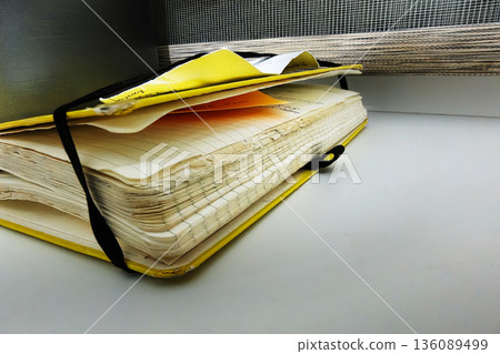 A yellow notebook sits on a table, with its pages slightly worn. Papers are placed on top, and sunlight comes through the window nearby in the afternoon 136089499